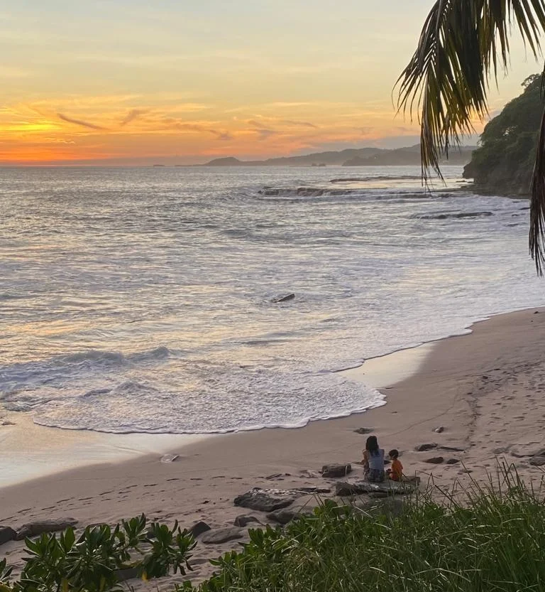 A peaceful sunset at Playa Rosada, Nicaragua, with a woman and one children sitting on the sand, watching the waves.