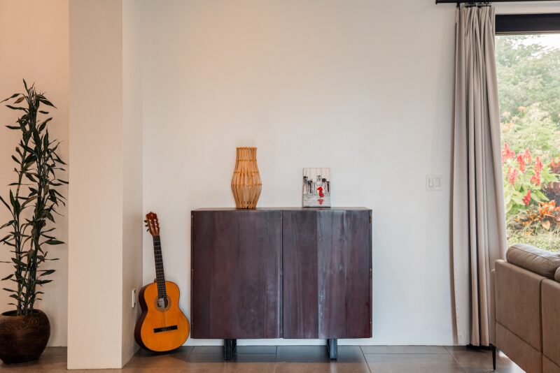 Minimalist living room corner with wooden cabinet, guitar, and indoor plant beside a curtained window.