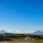 Panoramic view of a rural farm property with two volcanoes, open fields, trees, and small buildings in the distance.